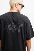 Ink Black | A man viewed from behind wearing an oversized black t-shirt with a tonal dark gray graphic of three surfers carrying surfboards across the back. He is also wearing black sunglasses and a gold chain necklace.