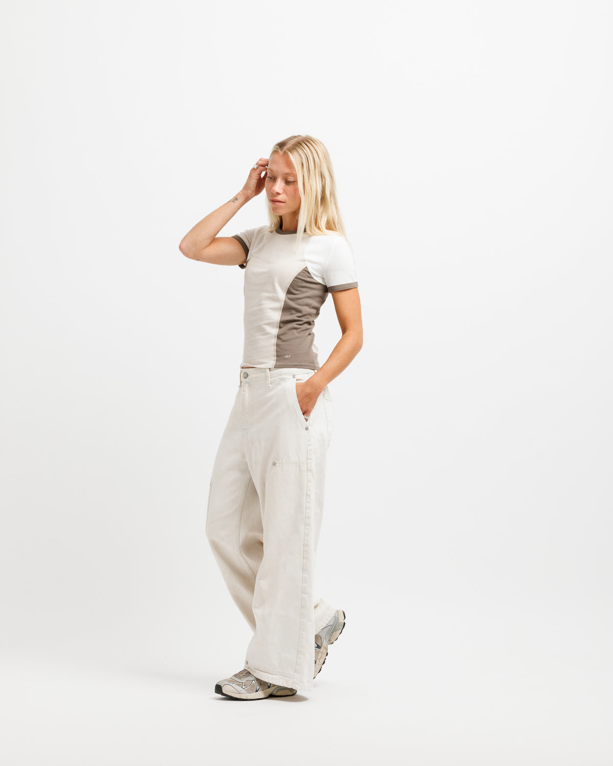 A woman modeling a short-sleeve t-shirt with a color-blocked design in off-white and taupe, paired with wide-leg off-white trousers and gray sneakers, against a plain white studio background.