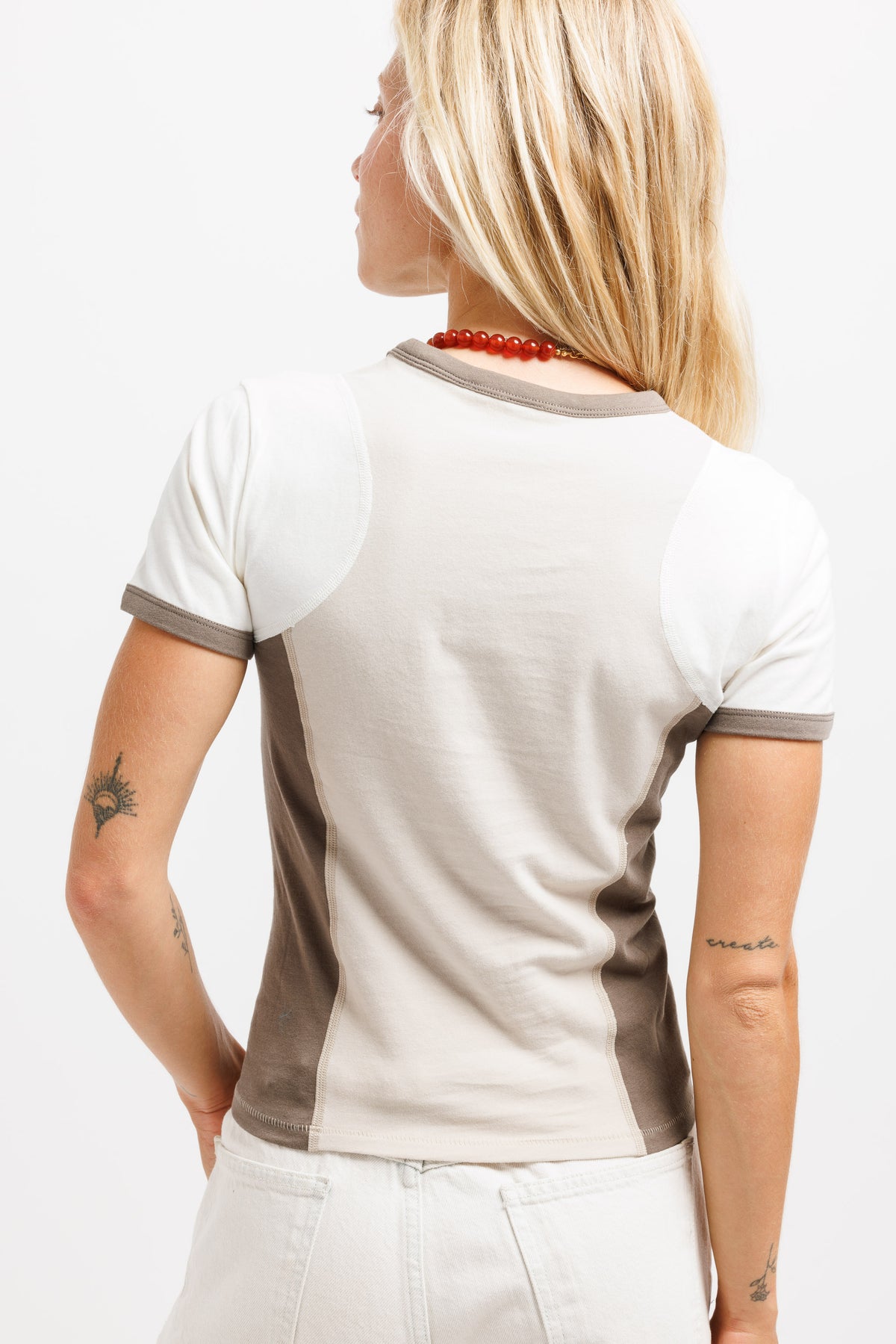 A woman shown from the back wearing a short-sleeve ringer t-shirt with a colorblock design in off-white and taupe, paired with white pants against a plain white background.