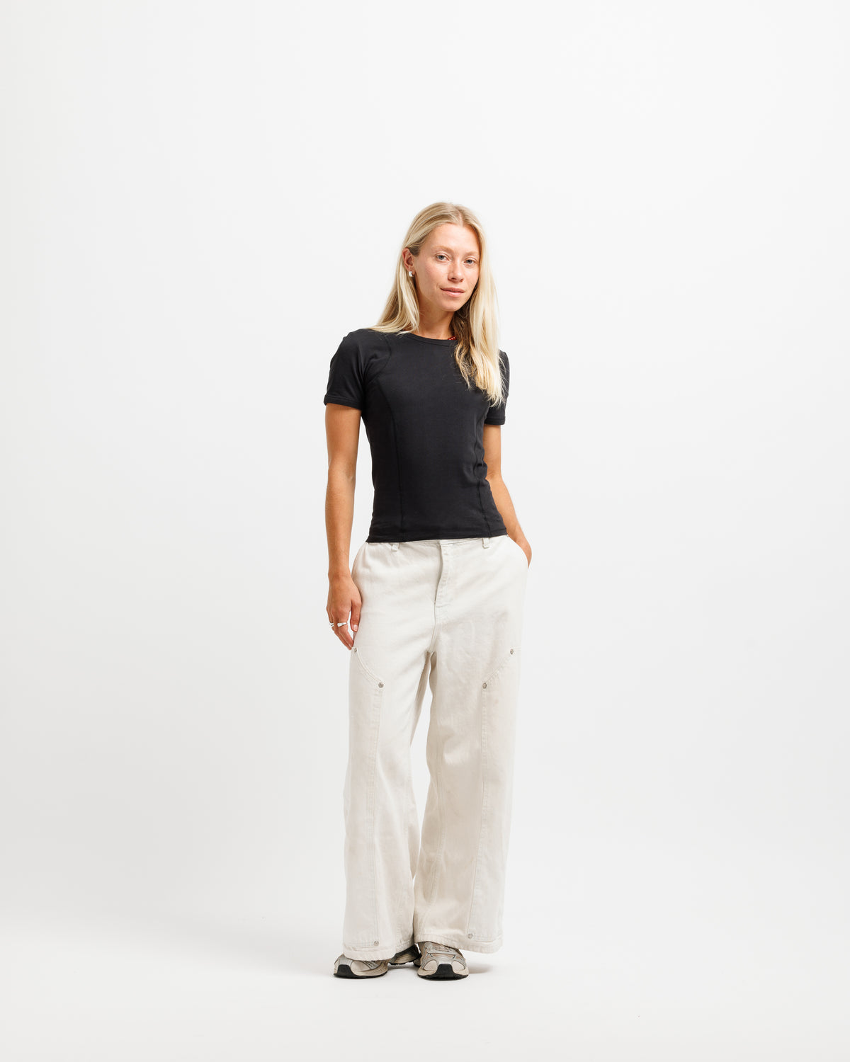 A woman stands against a plain white studio background wearing a fitted black short-sleeve top and loose off-white wide-leg pants with small dark embroidered details near the lower legs.