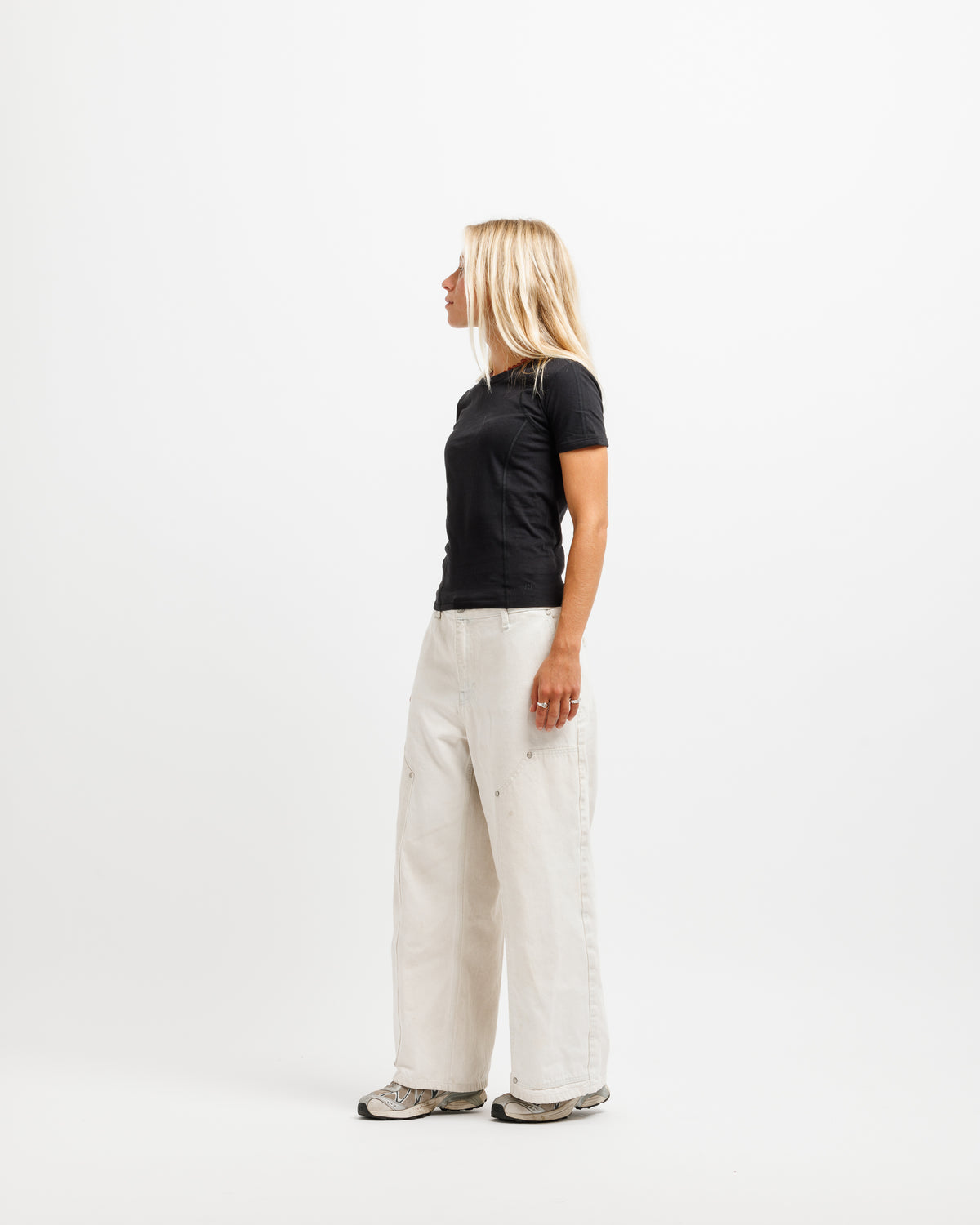 A woman stands in profile against a plain white studio background, wearing a black short-sleeve top, wide-leg off-white pants, and gray sneakers.
