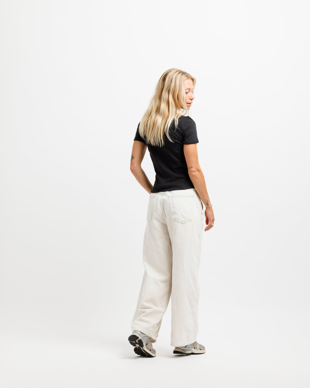 A blonde woman is shown from the back wearing a black short-sleeve t-shirt, off-white wide-leg linen pants, and gray sneakers, standing against a plain white studio background.