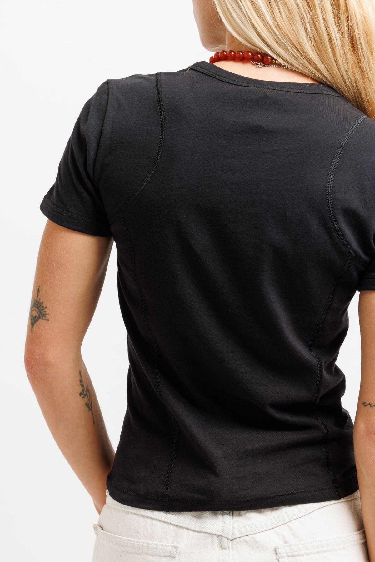 Rear view of a woman wearing a black short-sleeve crewneck t-shirt and white pants against a white studio background.