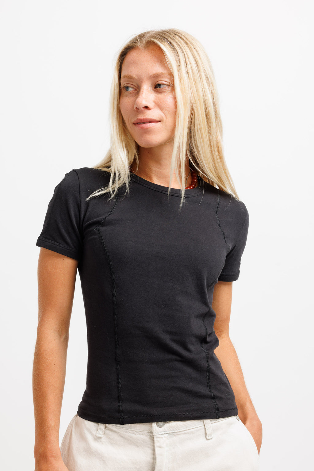 A blonde woman wearing a fitted black short-sleeve t-shirt and off-white pants, standing against a plain white studio background.