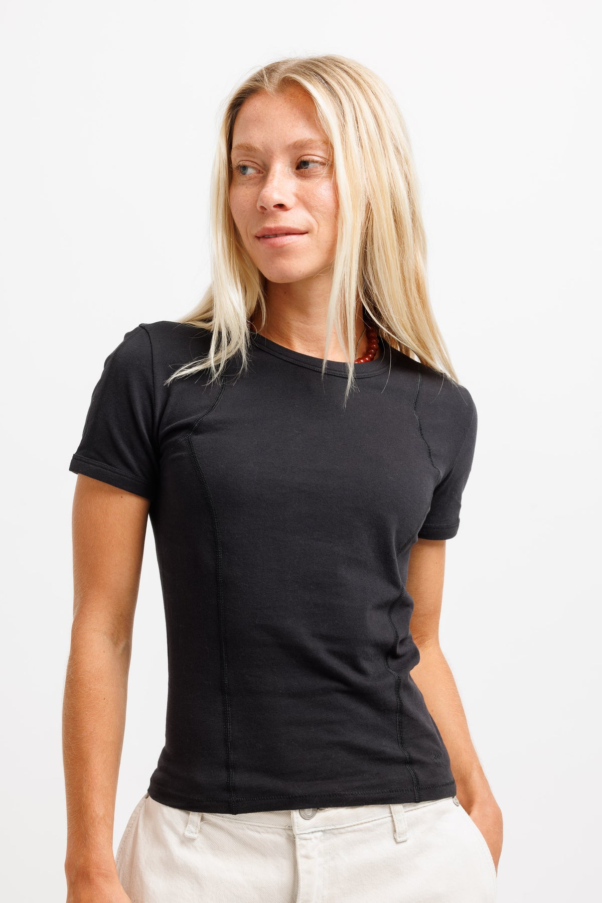 A blonde woman wearing a fitted black short-sleeve t-shirt and off-white pants, standing against a plain white studio background.