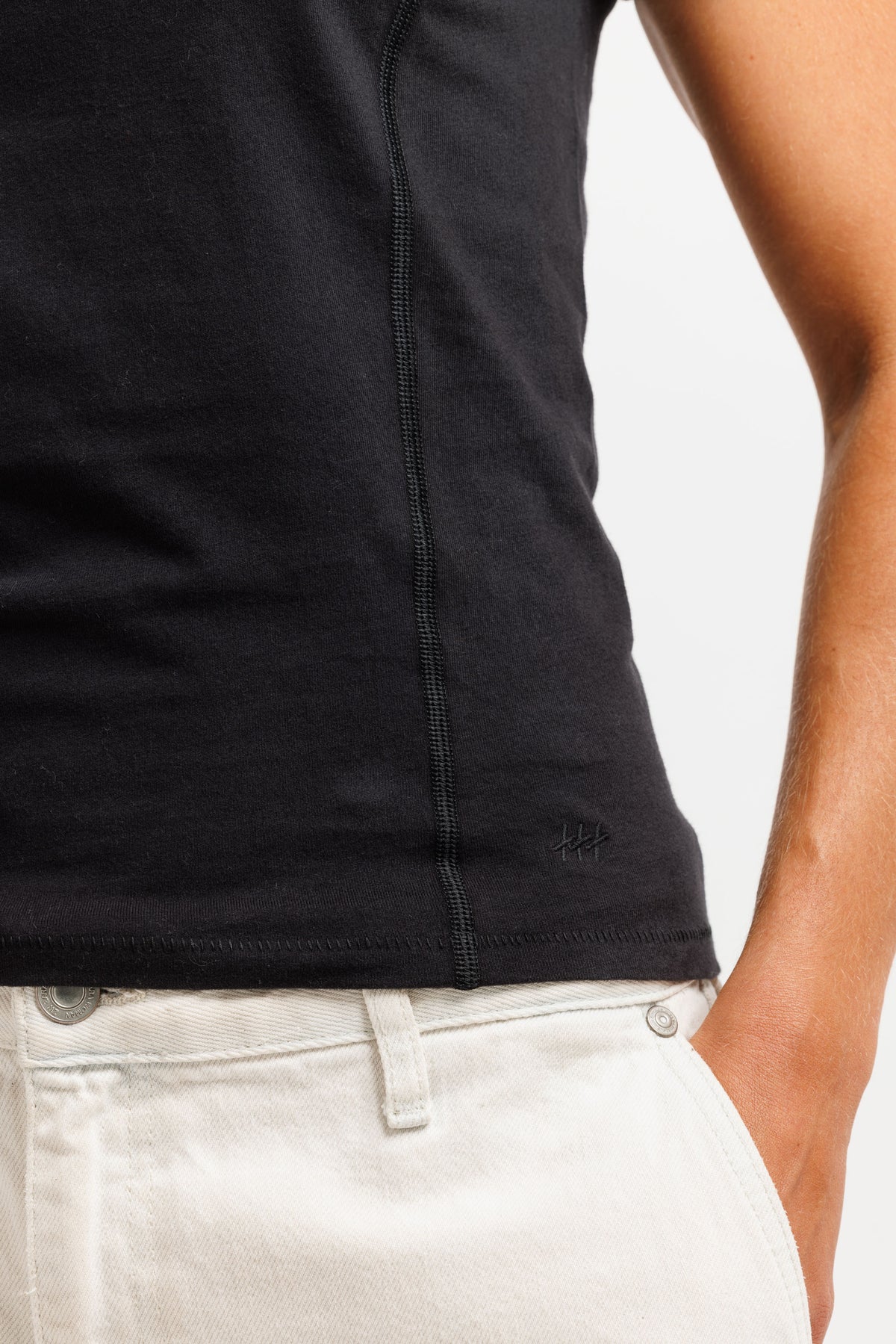 Close-up of a person wearing a black sleeveless top with a visible center seam and a small tonal logo near the lower right hem, paired with off-white pants against a light gray background.