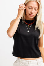Ink Black | A blonde woman wearing a black sleeveless crop top, layered silver necklaces, and a red beaded necklace, paired with light beige pants, against a plain white background.