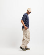 Deepwater Navy | On Model Full Side 2 A model with short blonde hair and sunglasses is shown in profile wearing a navy blue t-shirt, beige cargo pants, and dark sneakers against a plain white studio background.