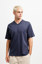 Deepwater Navy | On Model Front A man with platinum blonde hair wearing a navy blue v-neck t-shirt and beige pants, standing against a plain white studio background.