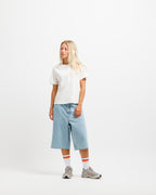 Cougar Milk | On Model Full Front A woman stands against a plain white studio background wearing a white short-sleeve t-shirt, light blue cropped denim shorts, white socks with red stripes, and gray sneakers.