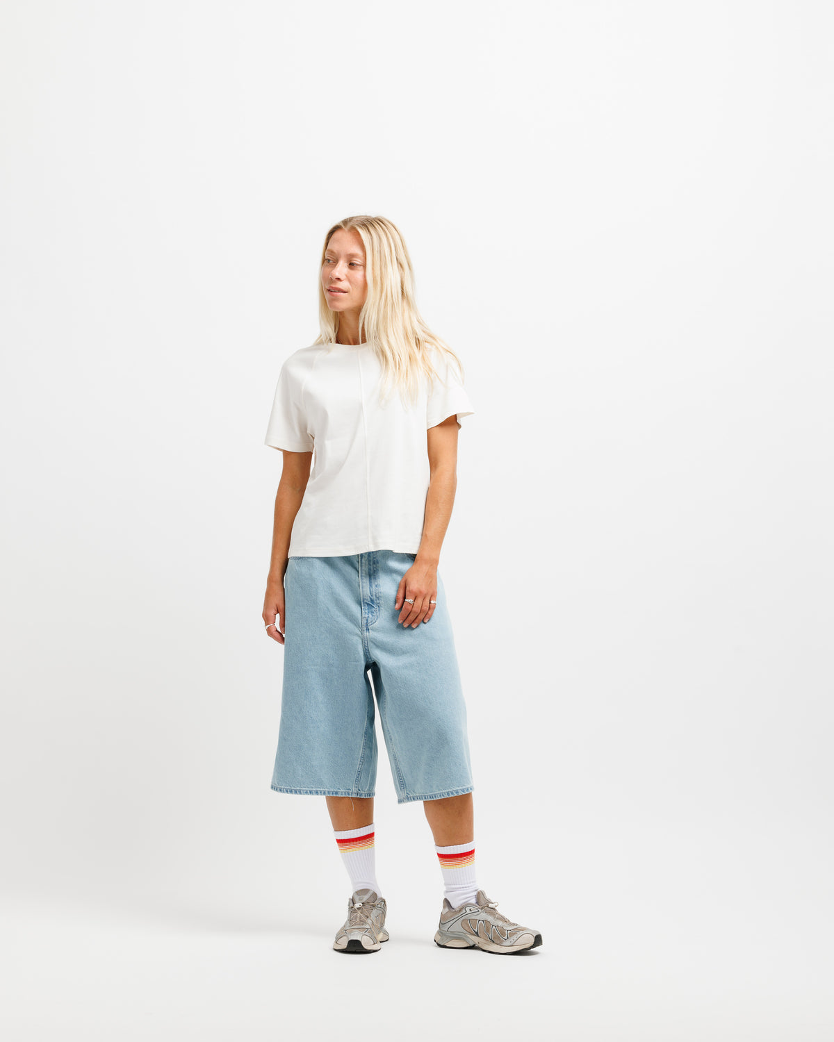 A woman stands against a plain white studio background wearing a white short-sleeve t-shirt, light blue cropped denim shorts, white socks with red stripes, and gray sneakers.