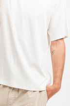 Cougar Milk | On Model Back Detail Close-up of a person wearing a plain white short-sleeve T-shirt and light beige pants, with the hand in a pocket and a small tattoo visible on the upper arm against a light neutral background.