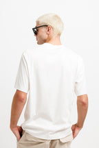 Cougar Milk | On Model Back A man with short blonde hair and black sunglasses, viewed from behind, wearing an oversized white short-sleeve t-shirt and beige pants against a plain white background.