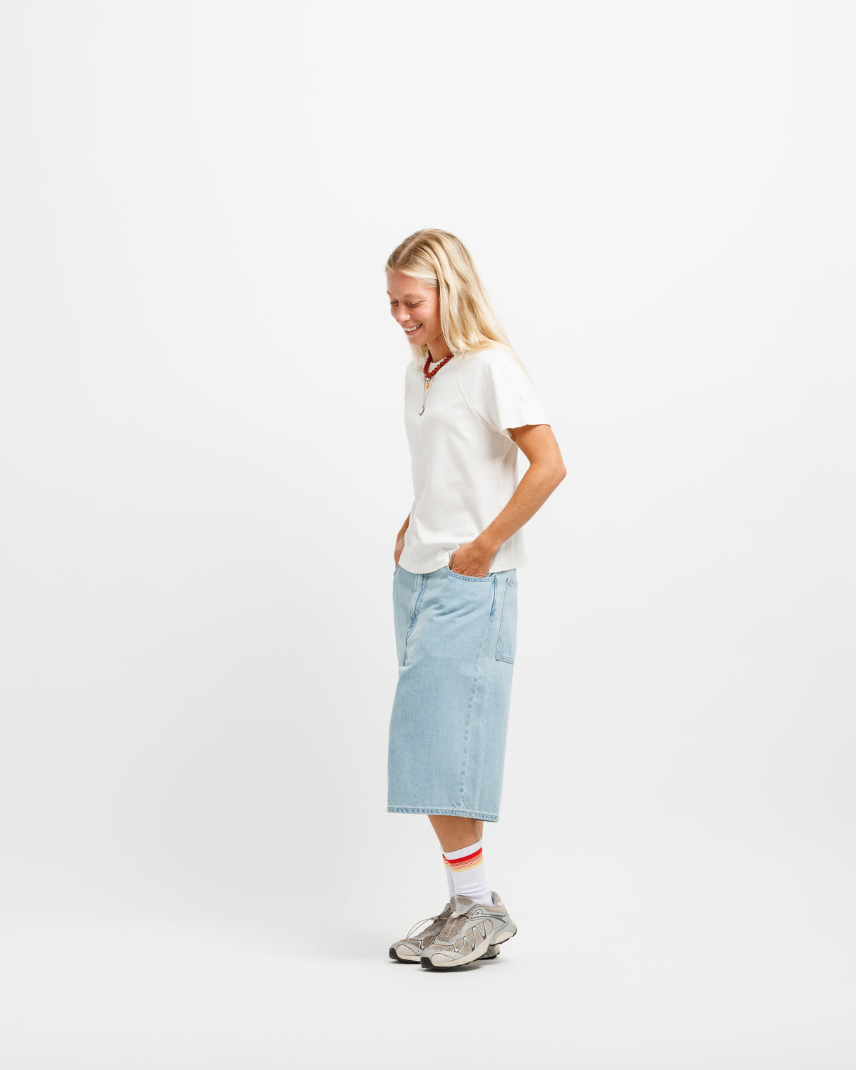 A woman stands in profile wearing a white short-sleeve crew, a light blue denim midi skirt, white socks with red stripes, and gray sneakers against a plain white studio background.