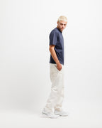 Deepwater Navy | On Model Full Side A model with platinum blonde hair stands in profile wearing a navy blue crewneck t-shirt, off-white relaxed-fit trousers, and white sneakers against a plain white studio background.