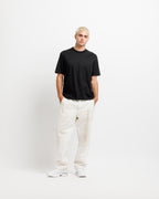 Ink Black | On Model Full Front A man with short blonde hair stands against a plain white studio background wearing a black short-sleeve crewneck t-shirt, off-white linen pants, and white sneakers.