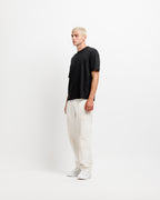 Ink Black | On Model Full Side A man stands in profile wearing a black short-sleeve t-shirt, off-white linen trousers, and white sneakers against a plain white studio background.