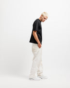 Ink Black | On Model Full Side 2 A model with platinum blonde hair is shown in profile wearing a black short-sleeve t-shirt, off-white textured trousers, and white sneakers against a plain white studio background.