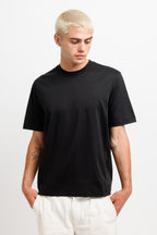 Ink Black | On Model Front A man with platinum blonde hair wearing a plain black short-sleeve crewneck t-shirt and off-white trousers, standing against a light gray studio background.