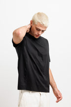 Ink Black | On Model Side A man with platinum blonde hair wearing a black short-sleeve t-shirt and off-white trousers, posing against a solid white studio background.