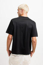Ink Black | On Model Back A man with platinum blonde hair is shown from the back wearing a black short-sleeve crewneck t-shirt and off-white trousers, photographed against a clean white studio background.
