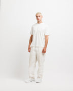 Cougar Milk | On Model Full Front A model with platinum blonde hair stands in a studio wearing a white short-sleeve t-shirt, off-white linen trousers, and white sneakers against a plain white background.