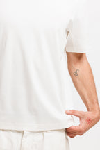 Cougar Milk | On Model Front Detail Close-up of a person wearing a plain white short-sleeve T-shirt and light-colored pants, with the hand pulling the hem slightly outward. A small heart tattoo is visible on the forearm against a clean white background.
