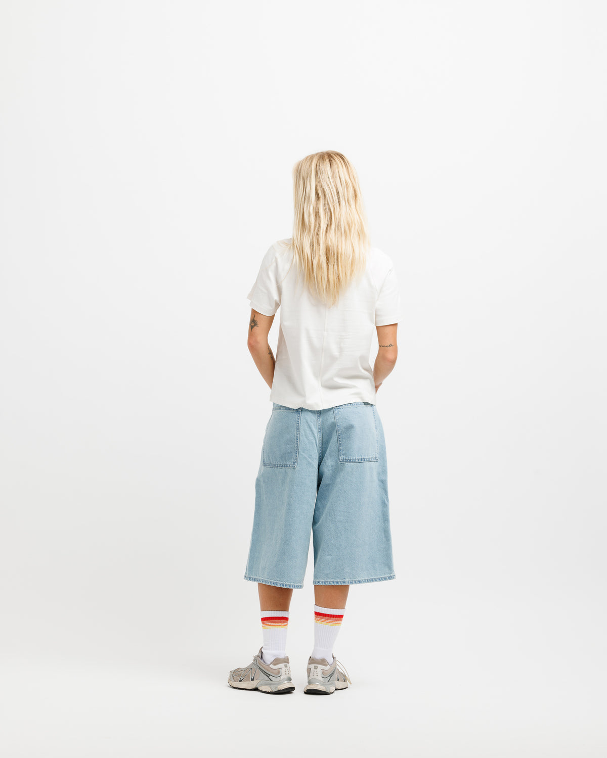 A model viewed from behind wearing a white t-shirt, light blue denim cropped pants, white socks with red stripes, and gray sneakers, set against a plain white studio background.