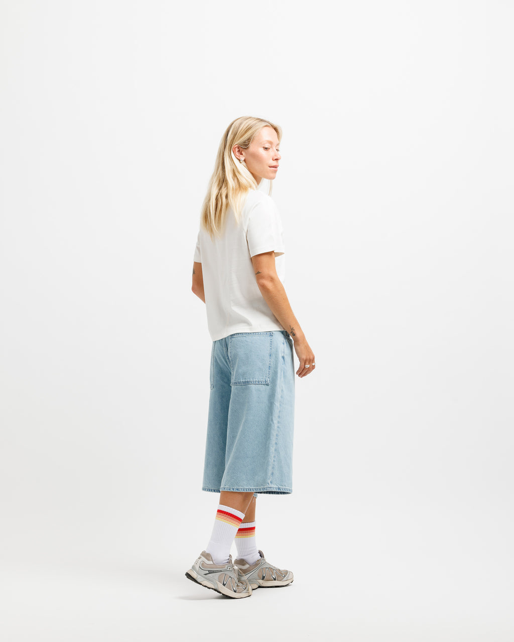 A woman stands in profile wearing a white short-sleeve T-shirt, light blue wide-leg denim shorts, white socks with red stripes, and gray sneakers against a clean white studio background.