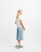 Cougar Milk | On Model Full Side A woman stands in profile wearing a white short-sleeve T-shirt, light blue wide-leg denim shorts, white socks with red stripes, and gray sneakers against a clean white studio background.
