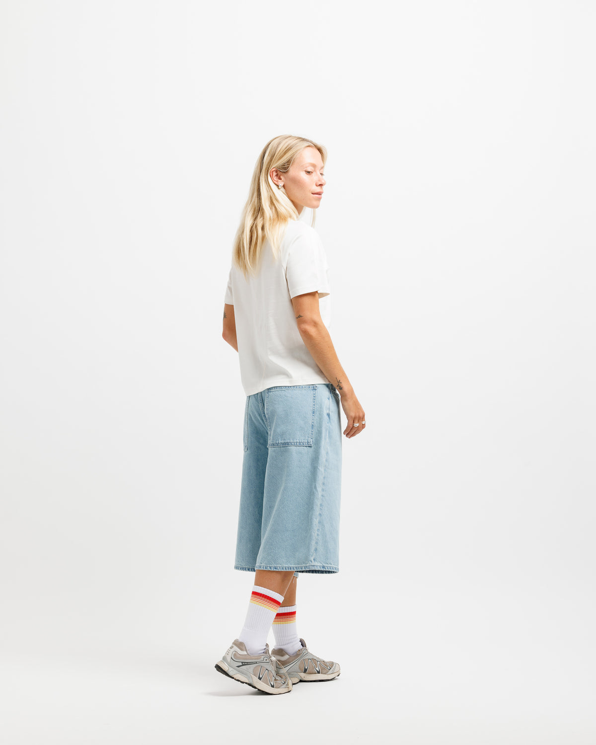 A woman stands in profile wearing a white short-sleeve T-shirt, light blue wide-leg denim shorts, white socks with red stripes, and gray sneakers against a clean white studio background.