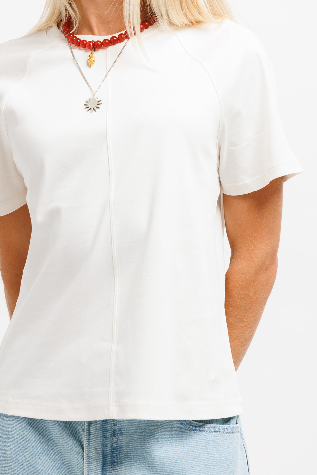 A close-up of a person wearing a white short-sleeve top with a split V-neckline, paired with light blue jeans and layered necklaces.