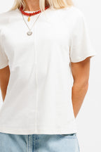 Cougar Milk | On Model Front Detail A close-up of a person wearing a white short-sleeve top with a split V-neckline, paired with light blue jeans and layered necklaces.