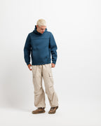 Stillwater Blue | On Model Full Front A man with platinum blonde hair stands in a studio wearing a dark teal-blue hooded fleece jacket, beige cargo pants, and brown hiking boots against a solid white background.