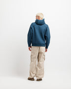 Stillwater Blue | On Model Full Back A model viewed from the back wearing a slate blue pullover hoodie and beige cargo pants against a plain white studio background.