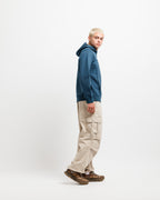 Stillwater Blue | On Model Full Side A model with platinum blonde hair stands in profile wearing a dark teal blue hoodie, beige cargo pants, and brown leather sneakers against a plain white studio background.