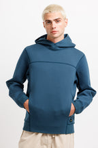 Stillwater Blue | On Model Front A man wearing a dark teal blue pullover hoodie with a unique asymmetrical chest seam and kangaroo pockets, paired with beige pants, against a plain white studio background.
