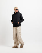 Ink Black | On Model Full Front A model with short blonde hair and sunglasses stands in a studio wearing a black pullover hoodie, beige cargo pants, and brown sandals against a plain white background.