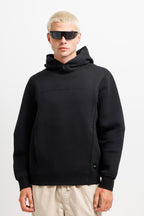 Ink Black | On Model Front A model with platinum blonde hair and sunglasses wearing a black pullover hoodie with a high folded collar and kangaroo pockets, paired with beige pants, against a plain white studio background.