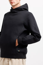 Ink Black | On Model Front Detail A close-up of a person wearing a black hooded sweatshirt with a cowl neck, long sleeves, and a kangaroo pocket, paired with light beige pants against a plain white background.