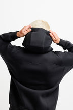 Ink Black | On Model Back A person viewed from behind wearing a black hoodie and pulling up the hood, with short blonde hair visible against a plain light gray studio background.