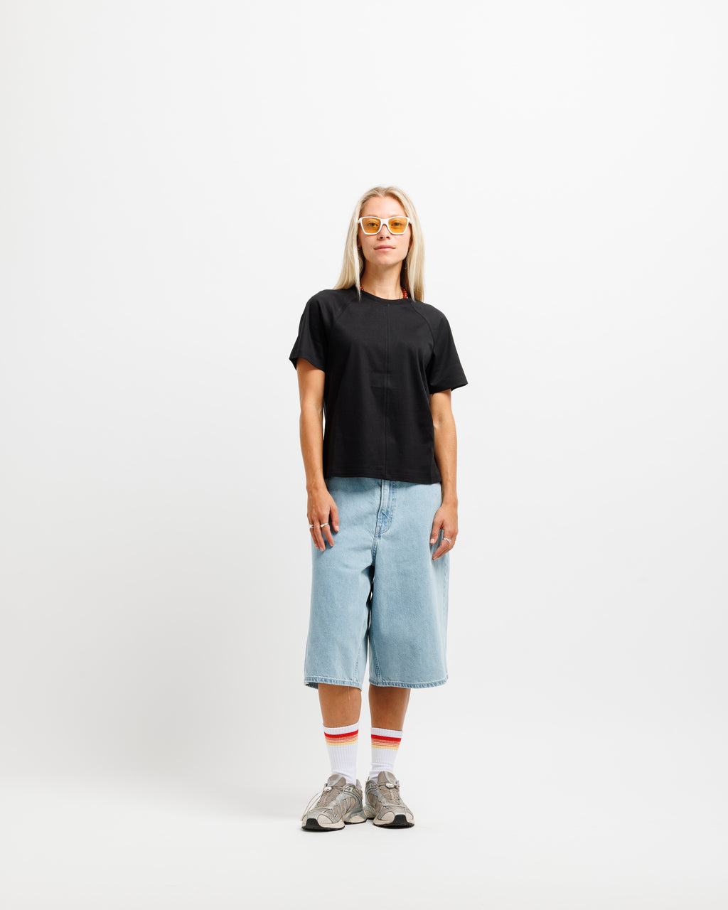 A woman stands against a plain white studio background wearing a black short-sleeve t-shirt, light-wash cropped denim shorts, white socks with red stripes, and gray sneakers.