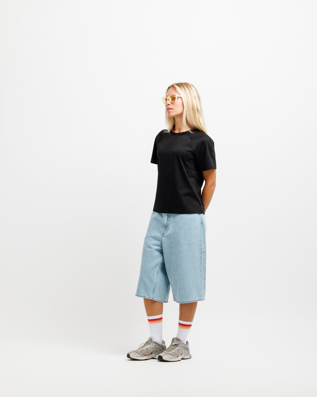 A woman stands in profile wearing a plain black short-sleeve T-shirt, light-wash denim shorts, white socks with red stripes, and gray sneakers against a clean white studio background.