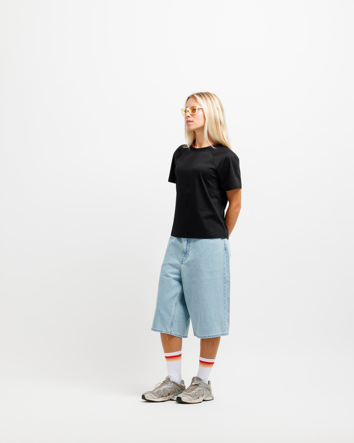 A woman stands in profile wearing a plain black short-sleeve T-shirt, light-wash denim shorts, white socks with red stripes, and gray sneakers against a clean white studio background.