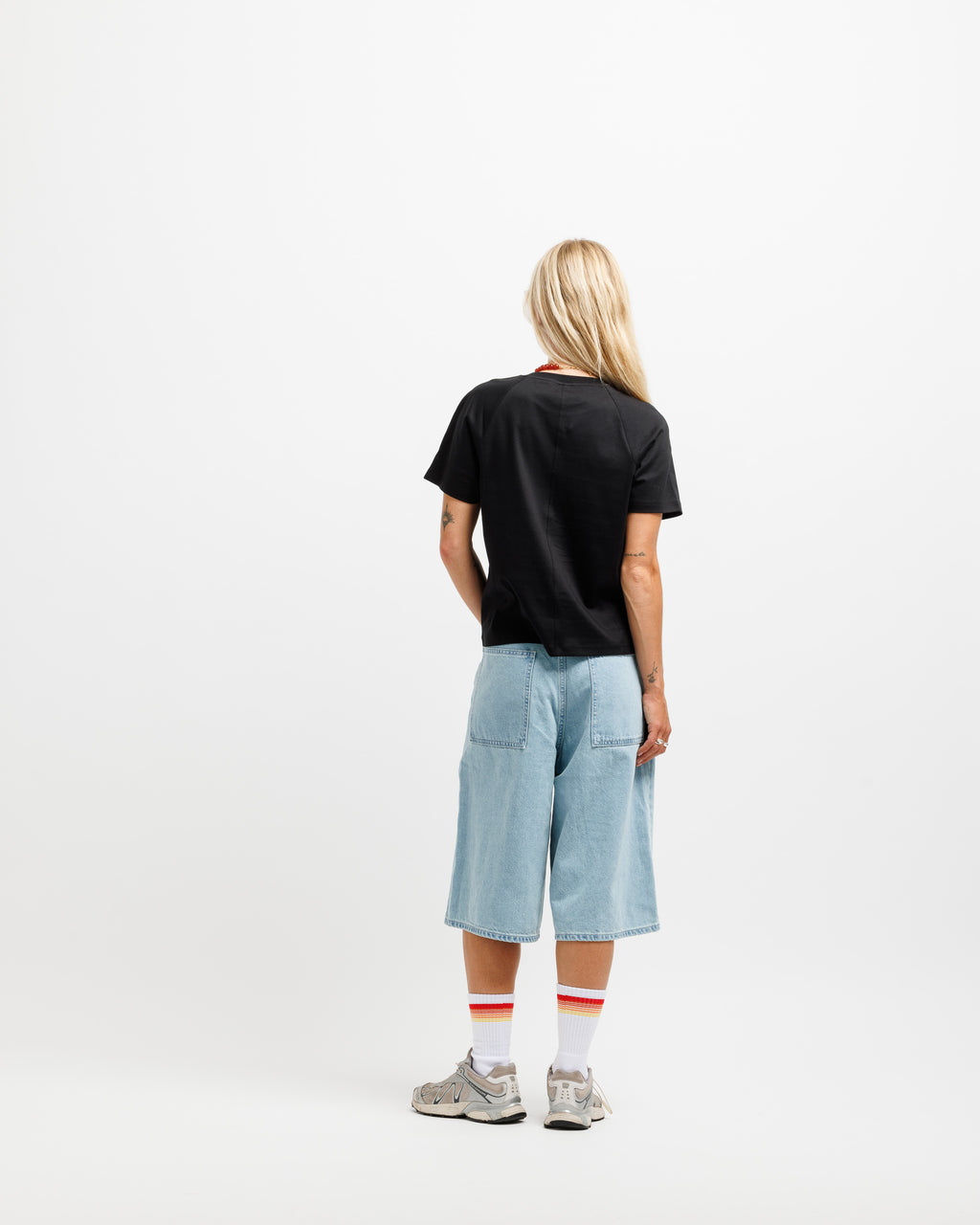 A model viewed from behind wearing a black t-shirt, light blue denim shorts, white socks with red stripes, and gray sneakers, standing against a plain white studio background.