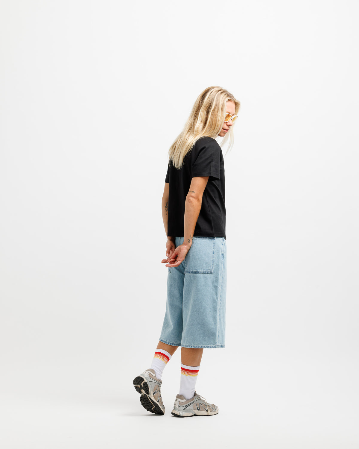 A woman stands in profile wearing a black short-sleeve T-shirt, light blue cropped denim shorts, white socks with red stripes, and gray sneakers against a plain white studio background.