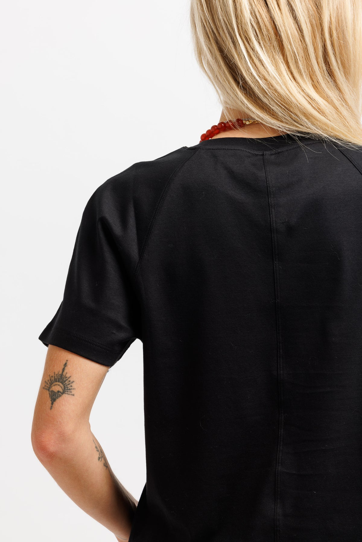 Rear view of a person wearing a black short-sleeve t-shirt, showing the back seam and a red beaded necklace against a plain white background.