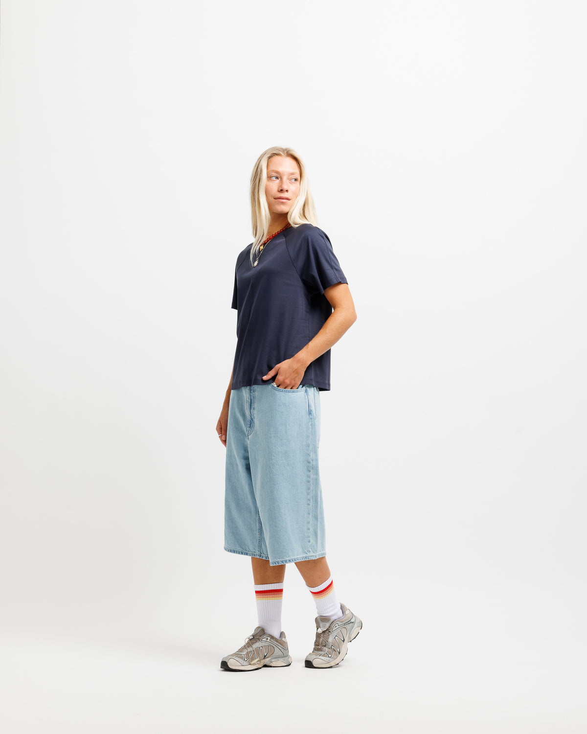 A woman modeling a dark navy blue t-shirt, light blue denim shorts, white socks with red stripes, and gray sneakers against a plain white studio background.