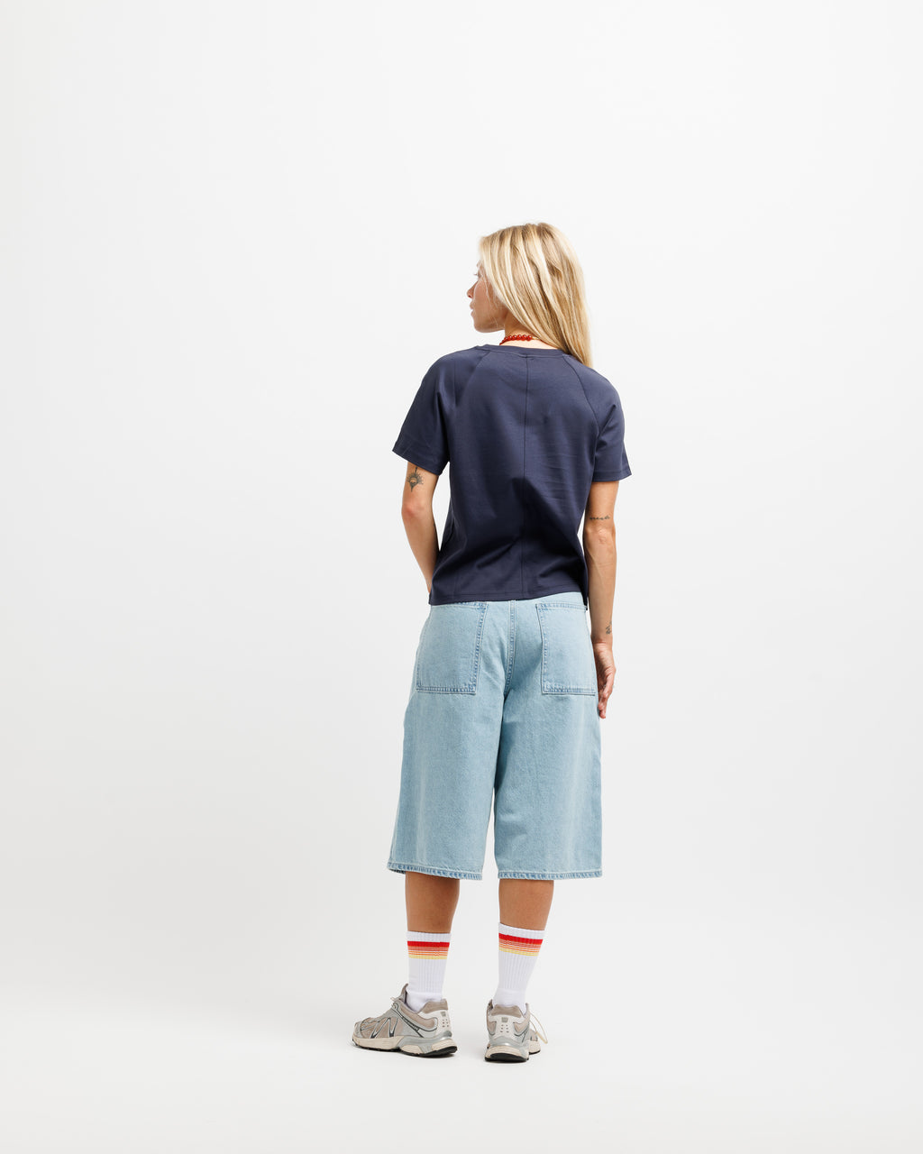 A woman is shown from behind wearing a navy blue t-shirt, light wash denim shorts, white socks with red stripes, and gray sneakers, standing against a plain white studio background.
